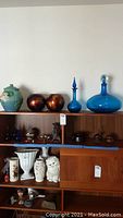 Photo shows Roseville cookie jar with blue-green glaze and raised water lily design, and two iridescent round vases, two blue glass decanters, and other items on wooden shelves.