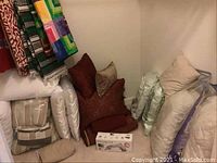 Assorted comforters, pillows in neutral tones, and boxed Rodents clothes steamer on floor in corner.