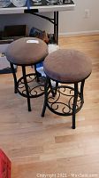 Pair of tan upholstered counter stools with black metal decorative bases showing full sides and seating area.
