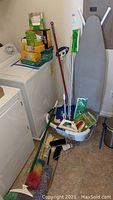 Wide image showing laundry area with washer, dryer, ironing board, broom, dustpan, mop, laundry basket, and cleaning supplies stacked on washer