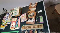 Photo of assorted children's books and wooden signs laid out on a table including a large rectangular 'WELCOME FRIENDS' sign and other smaller decorative signs.