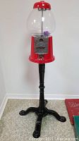 Full view of tall red standing gumball machine with black tripod base and clear globe on top.
