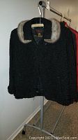 Full view of black short Persian lamb's wool coat with a fur trimmed collar, hanging on a rack.