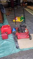 Overall view of red Landmark Mulch & Mow lawn mower with attached blue funnel, two red gas tanks, and garden soil bag on a tarp indoors
