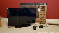 Front view of the Insignia 28 inch LED TV, original box behind TV, power adapter and remote in front.