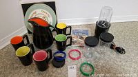 Full lot overview showing Magic Bullet blender base and jars, black pitcher, black mugs with colored interiors, decorative plates, and placemats.