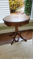 Full front and profile view of vintage round wooden side table with scalloped drawer and pedestal base standing on hardwood floor near window