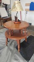 Front angle of the round two-tier wooden side table with the lamp on top showing turned legs and smooth finish.
