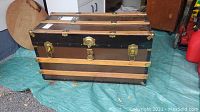 Front view of vintage steamer chest showing black body, wood straps, brass locks and latches, leather handles.