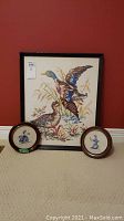 Photo showing all three needlepoint pieces including one large framed rectangular needlepoint of ducks and two smaller round framed needlepoints featuring duck designs.