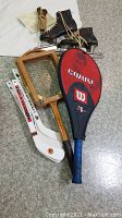 Photo showing a set of vintage sporting goods including a Wilson tennis racket with protective red cover, wooden framed vintage tennis racket, black leather speed skates, and hockey sticks laid out on floor.