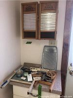 Wooden cabinet with two washboard doors closed, showing washboard surface and 'Dual Hand' labeling. Below are vintage irons, clothes pins, advertisement boxes, and small basket arranged on a counter.