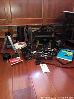 Photo showing vintage Nikkormat camera with worn leather case, Tamron zoom lens, additional Nikkormat lens, Carson binoculars in box, Biloret tripod and cleaning accessories laid on wood floor.