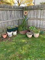 Wide shot of garden corner showing multiple plastic planters, buckets with stones, and metal sunflower decoration against wooden fence.