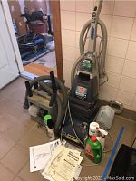 View of Hoover SteamVac B carpet cleaner with hoses and accessories in a room, accompanied by several bottles of cleaning solutions and user manual on floor.
