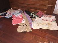 Image showing a variety of hand stitched pillow cases, aprons, and embroidered textile items laid out on a wooden floor.