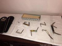 Photo of six vintage folding pocket knives laid out in open position alongside vintage scale in black case and decorative box liner