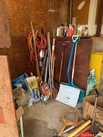 Wide view of corner showing snow shovels, broom, extension cords, bags of pellets, stakes, and maintenance bottles on upper wooden shelf.