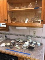 Overview of various porcelain and ceramic cups, saucers, bowls, and glassware displayed on countertop and cabinet shelf.