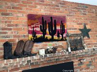 Wide view of the lot showing cowboy boots, hooked rug wall art with desert sunset and cactus design, whip, and western silhouette wall art frame against brick wall.