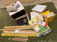 Photo showing multiple advertising yardsticks and rulers laid out with various printed company names and slogans, educational booklets, map box, and small yellow box with cards.