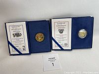 Two blue folders each containing a commemorative coin and a certificate of authenticity placed on a white surface.