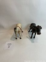 Two wooden Schoenhut horses, one white and one black, shown from the back side revealing tails and red straps.