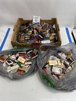 Large cardboard box filled with a variety of vintage matchbooks, showing many different designs and advertising logos.