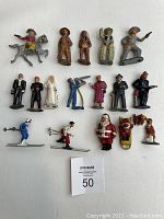 Top-down photo showing the full collection of 16 vintage lead figures arranged on a white surface, including cowboy, Native American, wedding couple, skiers, Santa Claus, and other characters.