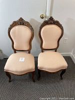 Pair of antique chairs with beige upholstery and carved wooden frames seen from the front.