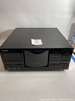 Front view of Pioneer Elite PD-F19 300 disc CD changer showing overall unit with disc tray in center, control panel with buttons and large dial, and a label on top of unit.