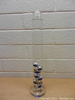 Full view of 17 inch tall glass Galileo thermometer with multiple blue, white, and gold colored spheres inside.
