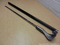 Black walking cane and long slender sword blade removed from cane shown on a tan surface.