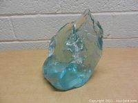 Solid light blue glass piece with jagged and uneven edges, displayed on a table against a white brick wall background.
