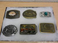Top-down view of six decorative metal belt buckles arranged on a white surface, showing varied designs including a stone centerpiece, eagles, rodeo scene, and logos.