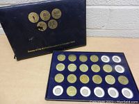 Blue presentation box open showing 20 gold-toned coins or medallions with five empty slots visible, arranged in a grid under a velvet-like lining.