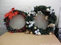 Two Christmas wreaths displayed leaning against a white brick wall on a brown table. Left wreath has red poinsettias and large red bow. Right wreath has white poinsettias and gold baubles with cherubs.