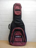 Front view of closed Road Runner guitar soft case standing against a white brick wall, showing black and maroon color scheme and front pockets with zippers.