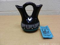 Front view of black ceramic Native American Kopa wedding vase with geometric painted band and turquoise accent. Blue tag present describing the item.