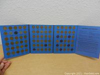 Blue tri-fold coin collector book holding numerous Lincoln pennies from 1941 to 1974, including slots with coins and empty slots indicating the set is almost complete.