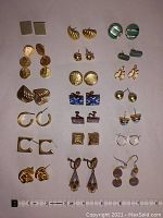 Overall view of 20 pairs of costume earrings arranged in five columns by four rows showing various styles including studs, hoops, drops, clip-ons, gold tone metal, enamel, and faux pearl designs. Included ruler for scale.