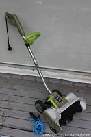 Electric snow shovel in a standing position showing handle, cord, wheels and snow intake.