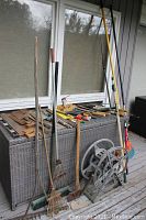 Overview of assorted garden tools including rakes, hoes, hand tools, extension pole, and hose reels against a deck box.
