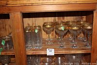 Wood cabinet shelf with three groups of glassware: six gold-trimmed wine glasses on the right, six tall glasses with gold trim in the middle, and six short glasses with gold trim on the left lower shelf.