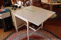 White IKEA drop leaf table shown in a room with other furniture. Table is partially folded and has four legs with a support bar.