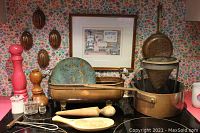 Photo showing various kitchen items on a stove against floral wallpaper including copper molders, a framed art print, copper warmed plate, metal bin, wooden grinders, wooden utensils, and glass salt and pepper shakers.