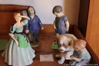 Four porcelain figurines displayed on wooden surface including a woman in a dress, two boys in coats and overalls, and a child petting a dog.