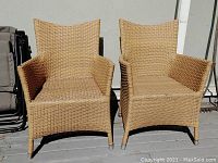 Front view of two matching natural-colored wicker chairs with armrests and high backs, set outdoors on a deck.