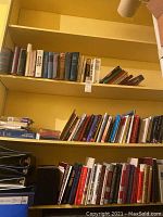 View of three shelves containing rows of reference and non-fiction books in various sizes and colors, mostly upright but some stacked horizontally. Bottom shelf shows several black binders.