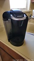 Black and silver Keurig single-serve coffee maker with attached water reservoir on left side, placed on counter near window.